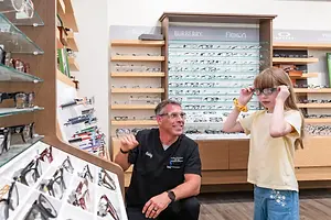 An adult man and a girl child are in an optical shop. The man is kneeling in front of the girl and appears to be explaining something to her. Behind them, several shelves are filled with eyeglasses, and the display case has a glass cover.