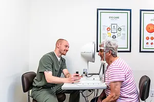 A man and a woman are inside a clinic room, where the man is sitting in front of a woman wearing a white headband. The man is adjusting the equipment in front of him. Behind them are two framed posters on the wall.