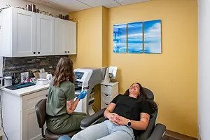 A woman sits in a chair with a smiling face while a woman in green stands beside her wearing gloves and a bracelet, with a cabinet, printer, and monitor nearby, and three blue sky and ocean paintings on the wall.
