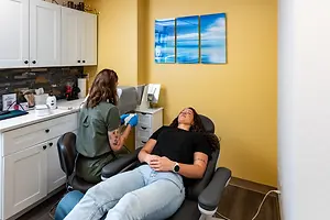 A woman is lying on a dental chair while the other woman is standing and holding a dental handpiece in front of the woman sitting. The woman sitting is wearing a watch and a bracelet. Behind them is a cabinet with drawers, a sink with a faucet, a roll of paper towels, a trash bin, and a power outlet on the wall. There are three paintings on the wall, and a power outlet is mounted on the wall.