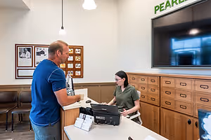 A man and a woman are interacting at a reception desk with a computer monitor and other items, and the wall is adorned with framed pictures, a monitor, and a sign reading Pearl.