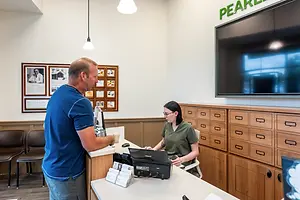 A man and a woman are interacting at a reception desk with a computer monitor and other items, and the wall is adorned with framed pictures, a monitor, and a sign reading Pearl.
