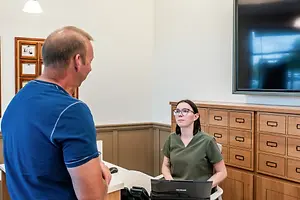 A man in a blue shirt is talking to a woman in a green shirt who is sitting at a desk with a laptop in front of her in an office setting.
