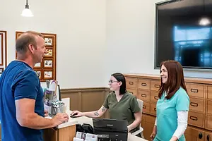 A man and woman are smiling and standing at the counter while a woman is sitting and using a computer monitor