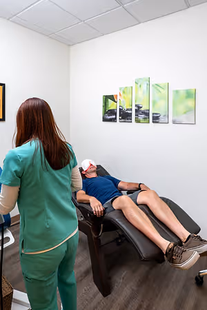 A woman in green scrubs stands near a man lying on a massage chair, wearing a blindfold and sneakers, while another woman in green scrubs looks on in a room with white walls, wooden floors, and framed artwork.