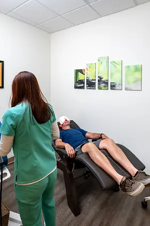 A woman in green scrubs stands near a man lying on a massage chair, wearing a blindfold and sneakers, while another woman in green scrubs looks on in a room with white walls, wooden floors, and framed artwork.