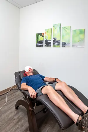 A man in a blue shirt and shorts lies on a massage chair with a blindfold, wires connected to his head, in a room with white walls and paintings of rocks and bamboo, possibly undergoing a light therapy session.