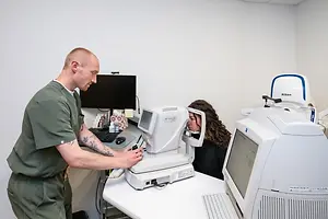 A man in a green shirt is adjusting an ophthalmoscope for a woman sitting in front of him in an eye exam room.