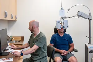 Two men are sitting in an examination room with an ophthalmoscope in the background, one wearing a watch and the other holding a remote control, both appear to be in a medical setting.