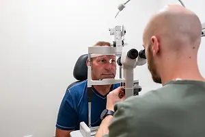 A man is sitting in a chair while a man wearing a green shirt is looking at his eyes with a microscope