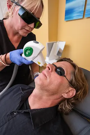 A woman wearing glasses is administering a laser treatment on a man's face in a clinic.