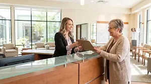 Receptionist assisting an elderly woman with a clipboard at the front desk in a modern medical facility.