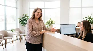 An older woman standing behind a reception desk talking to a younger woman sitting at a computer