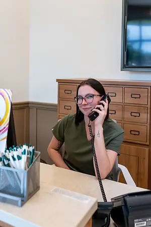 A woman wearing glasses and a green shirt is sitting on a chair and talking on the phone in an office room.