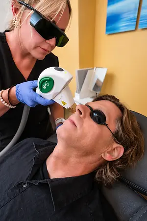 A woman is performing laser therapy on a man's face while he wears sunglasses