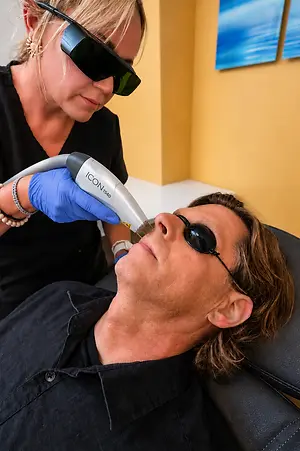 A woman is performing a laser treatment on a man's face while he lies on a chair wearing sunglasses and a bracelet.