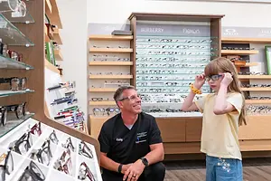 An adult male and young girl are inside an eyeglasses shop. The girl is holding a pair of glasses, and the adult male is sitting on a stool and smiling. Behind them are shelves with glasses, and a signboard with the word Burberry is mounted on the wall.