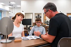 A man and a woman are talking to a boy sitting at a desk with a computer monitor in the background.