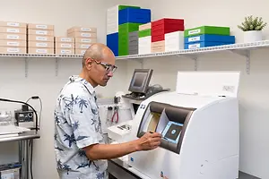 An adult male wearing glasses is standing in front of a machine while looking at a monitor in a lab.