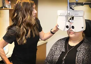A woman with long brown hair in a black shirt is adjusting an ophthalmoscope on a woman with leopard print clothing in an indoor setting.