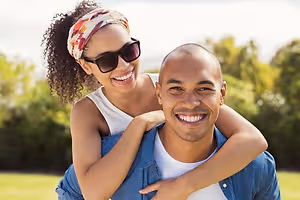 A man is giving a piggyback ride to a woman who is smiling and wearing sunglasses and a headband