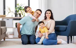 A family with a baby and a girl on the father's shoulders and the mother carrying a baby in her arms sitting on the floor in the living room