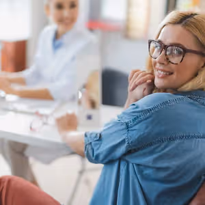 A woman wearing glasses and a blue shirt is sitting at a desk, smiling while looking at something. Another woman in a white lab coat is sitting in front of her, working on a computer.