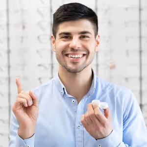 A smiling man in a blue shirt holds a pair of contact lenses in his hands