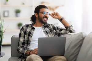 A man sitting on a couch, smiling, and adjusting his glasses while working on a laptop.