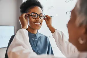 A smiling young woman trying on glasses at an optical store with a blurred background of other glasses on display.