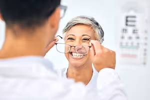 A woman with short gray hair smiles while looking into a mirror as a man in a white shirt adjusts her glasses.