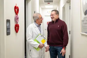 Two men stand in a hallway, one wearing a white lab coat, the other in a maroon sweater, both smiling and facing each other, one holding a folder, near a door with a red heart decoration, a sign, and a light on the ceiling