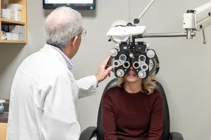 An elderly man, probably an eye doctor, is adjusting the eye machine of a woman who is sitting on a chair.