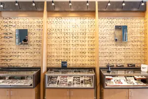 A room with wooden cabinets and glass displays showcasing eyeglasses, with a man standing behind the displays and lamps mounted on the ceiling.