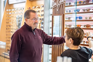 An older man in a maroon sweater and glasses is standing behind a glass display case, looking at sunglasses with an older woman wearing a black shirt and bracelet. Behind them, more sunglasses are displayed on shelves.
