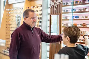 An older man in a maroon sweater and glasses is standing behind a glass display case, looking at sunglasses with an older woman wearing a black shirt and bracelet. Behind them, more sunglasses are displayed on shelves.