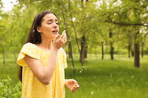A woman standing in a grassy field with trees behind her, she is blowing bubbles with her mouth and her eyes are closed. She is wearing a yellow dress with a white pattern.