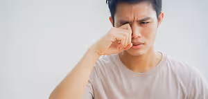 A man is rubbing his eye with his hand and seems to be in pain. He is wearing a white t-shirt. Behind him is a white wall. He is probably in a room with white walls and a white ceiling. He seems to be in a hospital or clinic.