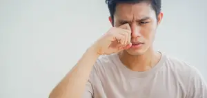 A man is rubbing his eye with his hand and seems to be in pain. He is wearing a white t-shirt. Behind him is a white wall. He is probably in a room with white walls and a white ceiling. He seems to be in a hospital or clinic.