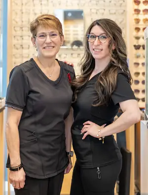 Two women in black uniforms standing and smiling in front of a wall with eyeglass frames