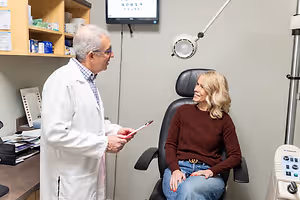 An elderly man in a white coat is talking to a woman in a chair in a clinic