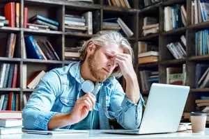 A man with a beard wearing headphones is sitting at a desk with a laptop and a cup of coffee. He is looking down and covering his face with his hand.