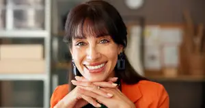 A smiling woman in an orange blazer poses for a photograph in an office setting.