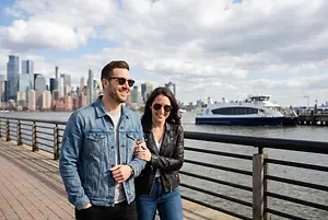 A man and woman are standing on a pier in front of a city skyline with a ferry boat in the background.