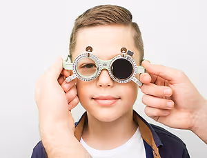A young boy wearing vision testing glasses is having his vision tested by an adult wearing a white shirt.