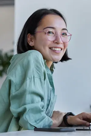 A woman with glasses is smiling and typing on a laptop.