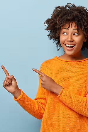 A young woman with curly hair, wearing an orange sweater, is smiling and pointing her index fingers at an unseen object in front of her.