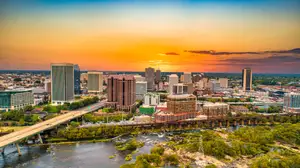 An aerial view of the city skyline at sunset, featuring tall buildings, a river, and a bridge with trees and greenery on the sides.