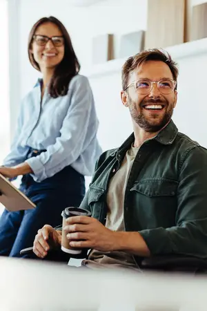 A man with a cup of coffee smiling and a woman with glasses in the background