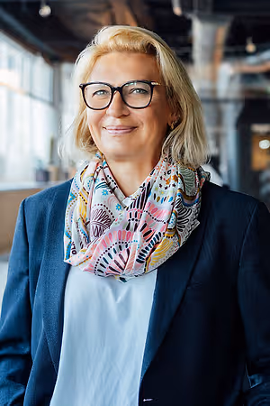 A smiling woman with blonde hair and glasses standing in a modern office with white walls and glass windows. She is wearing a blue blazer, a white shirt, and a colorful scarf around her neck. Behind her, there is a white shelf with some items on it.